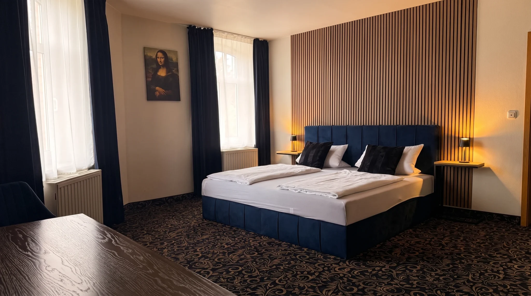 Doppelzimmer im Hotel In Amberg mit blauem Polsterbett vor Holzlamellenwand, Nachttischleuchten eingeschaltet